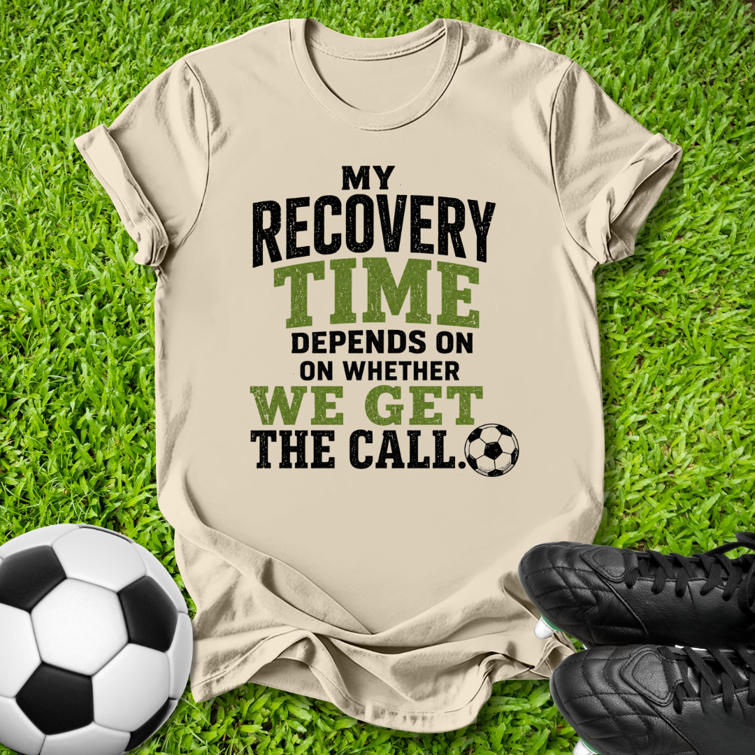 A beige graphic t-shirt laid flat on green grass, featuring the text 'MY RECOVERY TIME DEPENDS ON WHETHER WE GET THE CALL.' in black and green lettering, accompanied by a soccer ball and black cleats as props.
