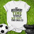A white graphic t-shirt laid flat on green grass, featuring the text 'MY RECOVERY TIME DEPENDS ON WHETHER WE GET THE CALL' in black and green lettering, accompanied by a soccer ball and black cleats.