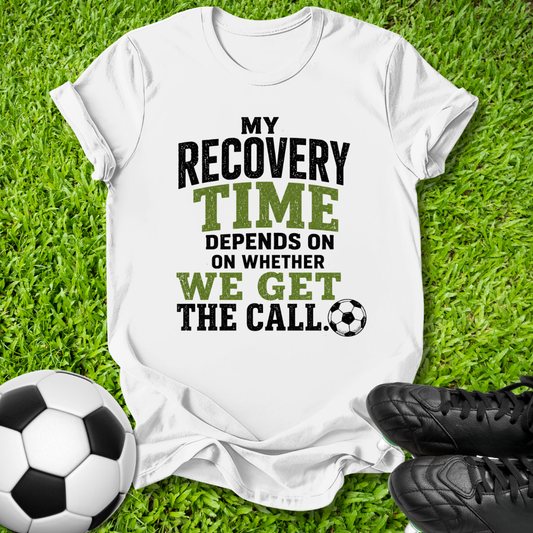 A white graphic t-shirt laid flat on green grass, featuring the text 'MY RECOVERY TIME DEPENDS ON WHETHER WE GET THE CALL' in black and green lettering, accompanied by a soccer ball and black cleats.
