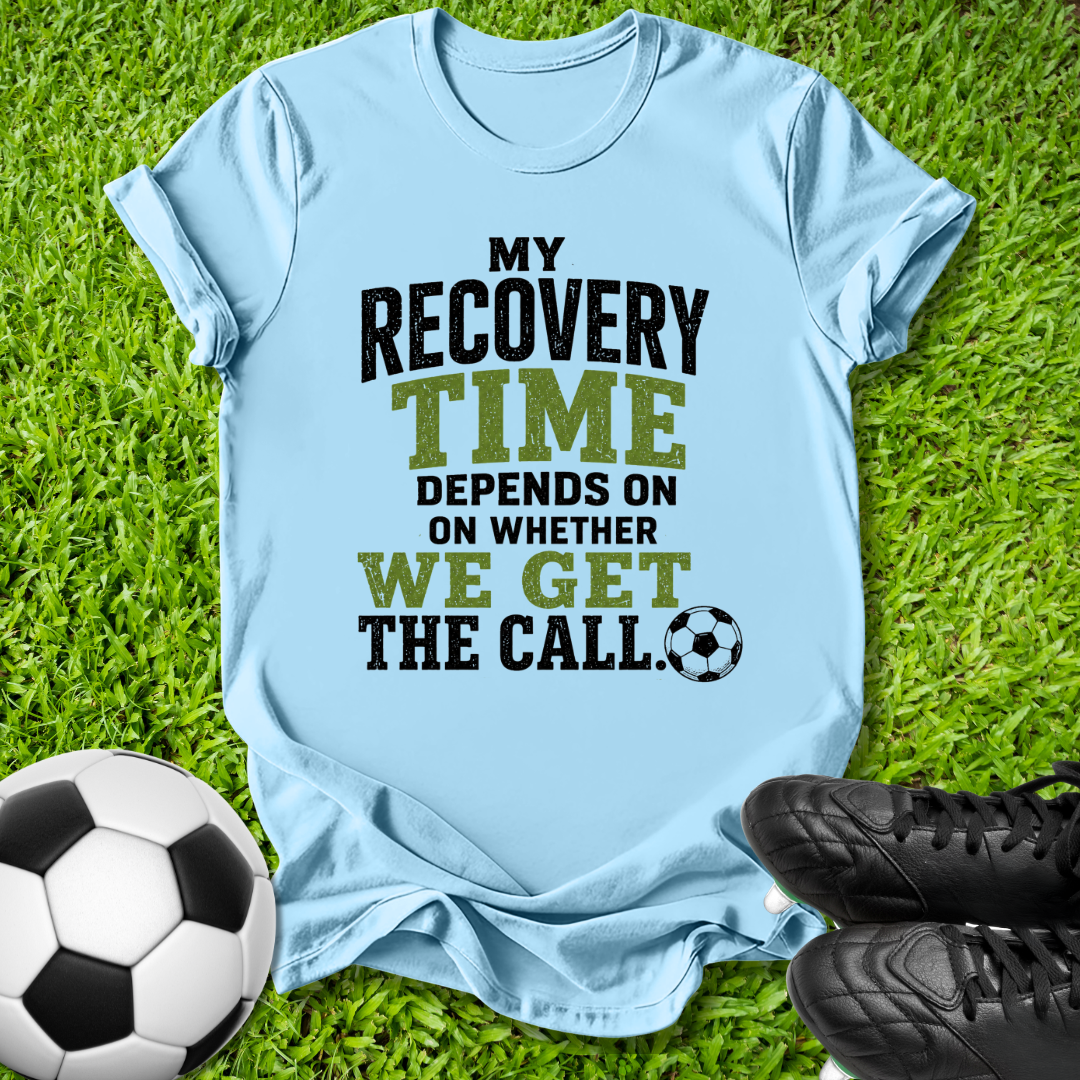 A light blue graphic t-shirt laid flat on green grass, featuring the text 'MY RECOVERY TIME DEPENDS ON ON WHETHER WE GET THE CALL.' in black and olive green lettering, accompanied by a soccer ball and black cleats.