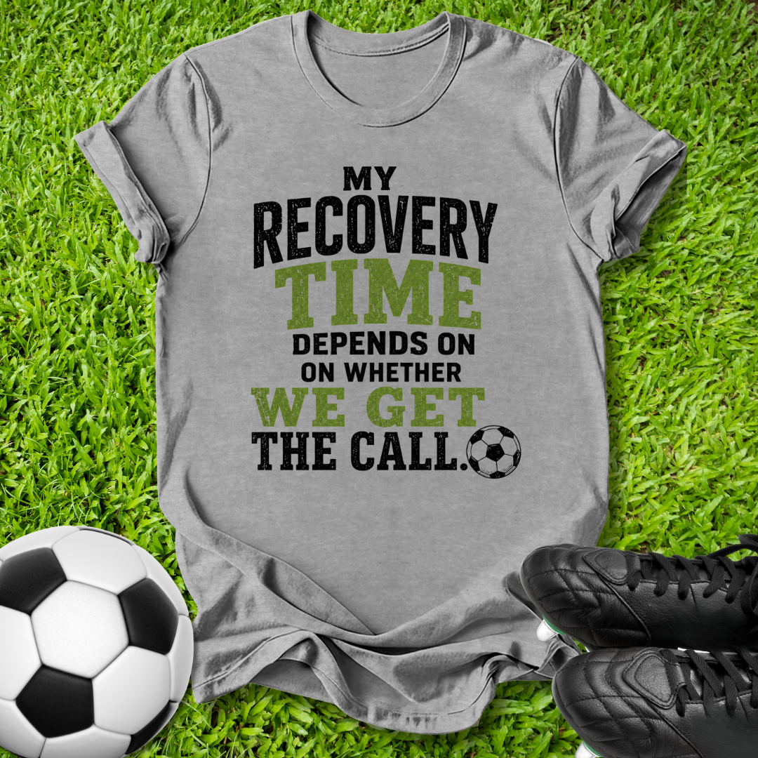 A heather gray graphic t-shirt laid flat on green grass, featuring the text 'MY RECOVERY TIME DEPENDS ON WHETHER WE GET THE CALL.' in black and olive green lettering, accompanied by a soccer ball and black cleats.
