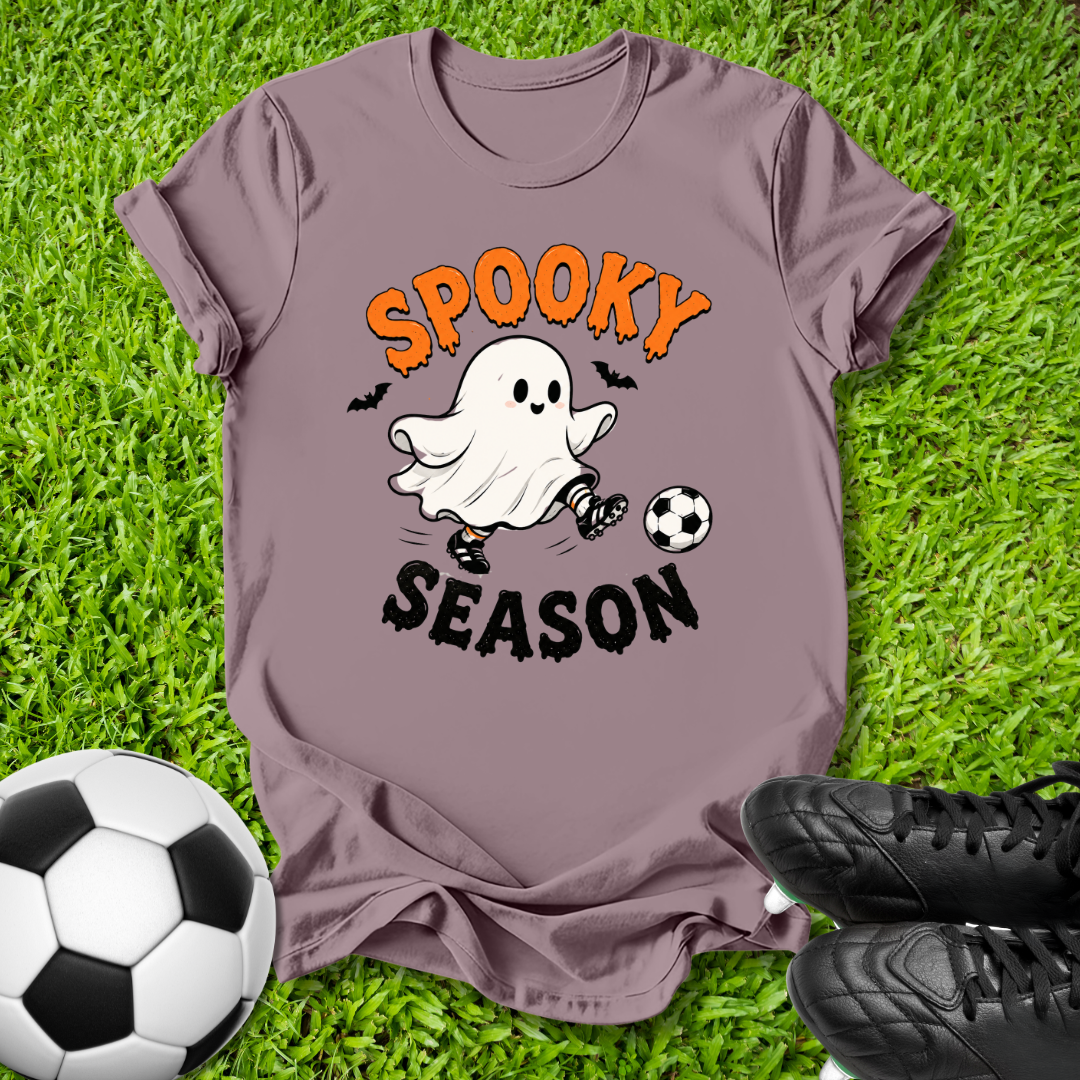 A mauve short-sleeve graphic T-shirt laid flat on green grass, featuring a cartoon ghost kicking a soccer ball with the words 'SPOOKY SEASON' in orange and black. A soccer ball and black cleats are placed around it as styling props.