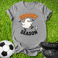 A heather gray graphic t-shirt laid flat on green grass, featuring a cartoon ghost kicking a soccer ball with the text 'SPOOKY SEASON' in orange and black. A soccer ball and black cleats are styled around the shirt.