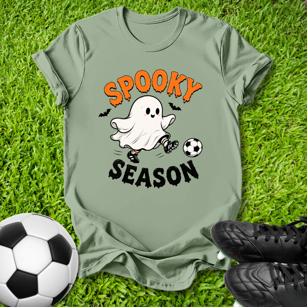 An olive green graphic t-shirt laid flat on green grass, featuring a cartoon ghost kicking a soccer ball with the text 'SPOOKY SEASON' in orange and black. A soccer ball and black cleats are placed around it as props.