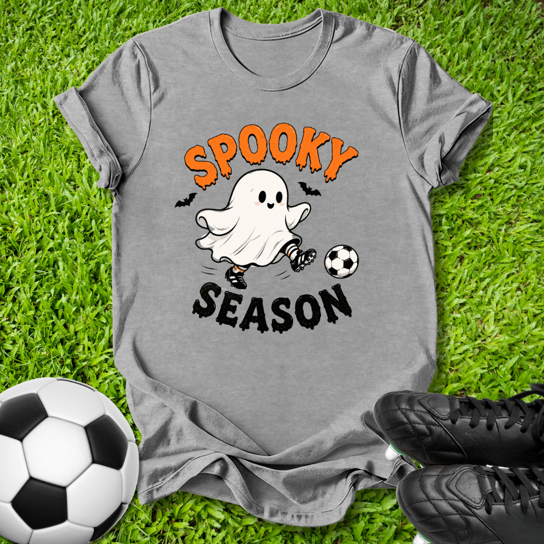 A heather gray graphic t-shirt laid flat on green grass, featuring a cartoon ghost kicking a soccer ball with the text 'SPOOKY SEASON' in orange and black. A soccer ball and black cleats are styled around the shirt.