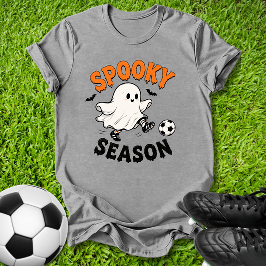 A heather gray graphic t-shirt laid flat on green grass, featuring a cartoon ghost kicking a soccer ball with the text 'SPOOKY SEASON' in orange and black. A soccer ball and black cleats are styled around the shirt.