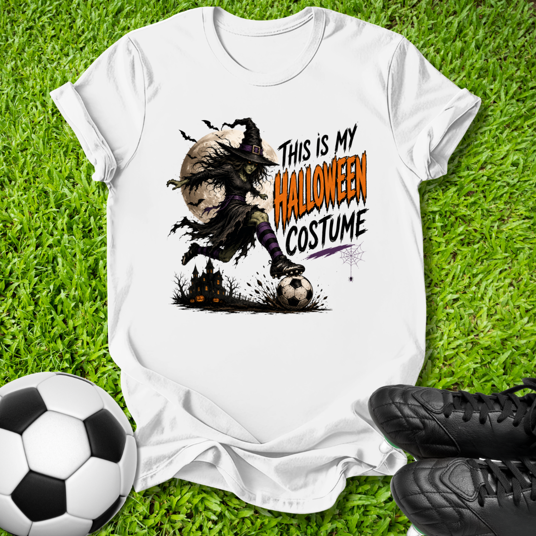 A white graphic T-shirt laid flat on green grass, with a soccer ball and black cleats around it. The shirt features a Halloween-themed illustration of a witch kicking a soccer ball, with the text 'THIS IS MY HALLOWEEN COSTUME' printed on the front.