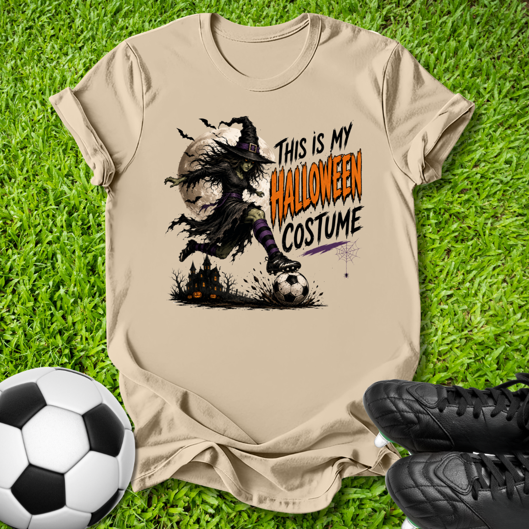 A beige graphic t-shirt laid flat on green grass, featuring a witch in a costume kicking a soccer ball with the text 'THIS IS MY HALLOWEEN COSTUME'. A soccer ball is on the left and black cleats are on the right.