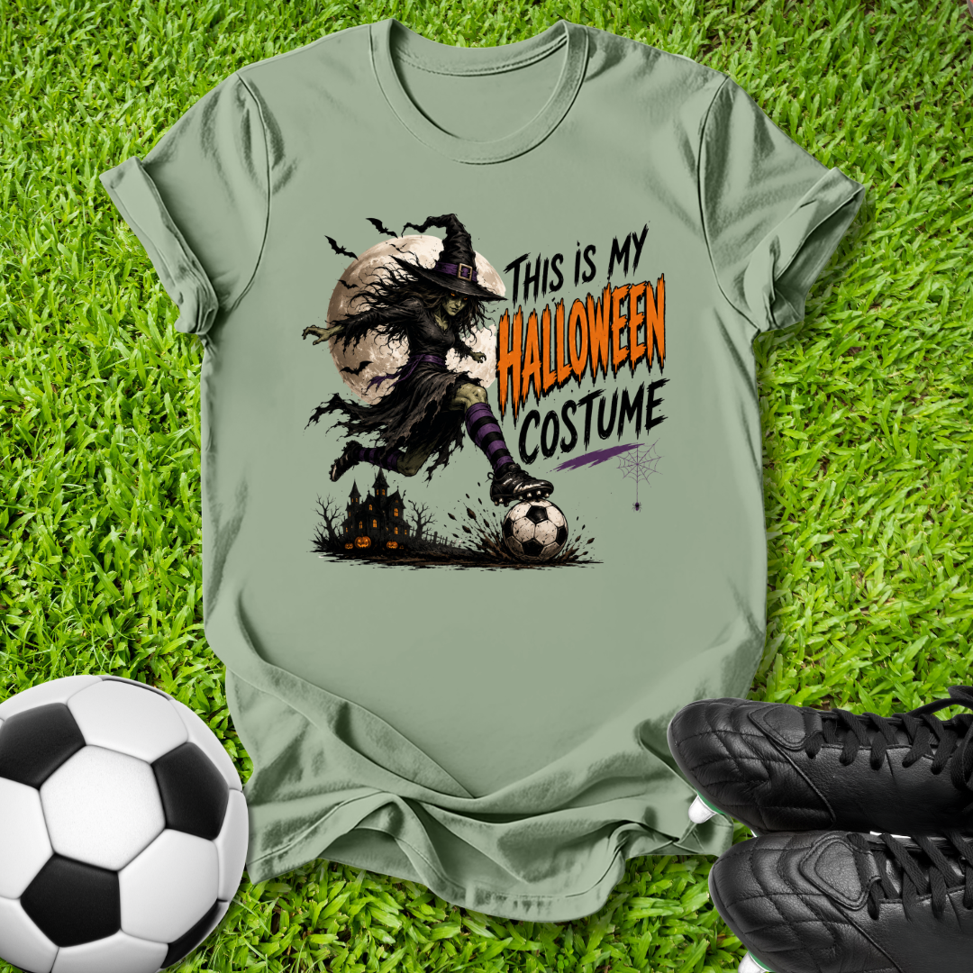 An olive green graphic t-shirt laid flat on green grass, styled with a soccer ball and black cleats. The t-shirt features a Halloween-themed graphic of a witch running with a pumpkin and the text 'THIS IS MY HALLOWEEN COSTUME'.