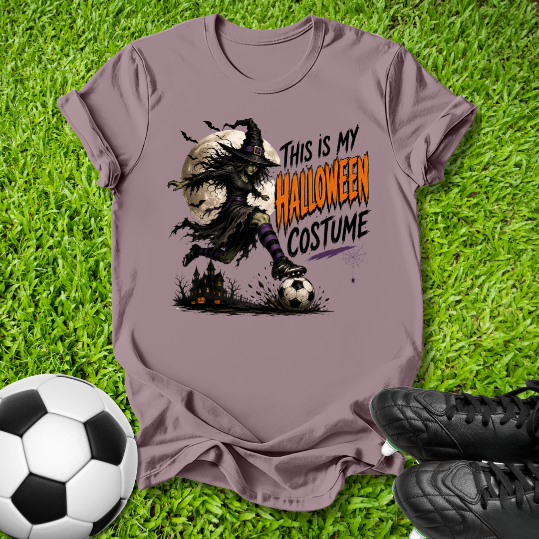A mauve-purple graphic t-shirt laid flat on green grass, featuring a spooky witch character in a soccer stance with the text 'THIS IS MY HALLOWEEN COSTUME'. A soccer ball and black cleats are placed around it as props.
