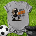 A heather gray graphic t-shirt laid flat on green grass, featuring a Halloween-themed illustration of a zombie-like figure in a costume kicking a soccer ball, with the text 'THIS IS MY HALLOWEEN COSTUME' in black and orange. A soccer ball and black cleats are placed around it as props.