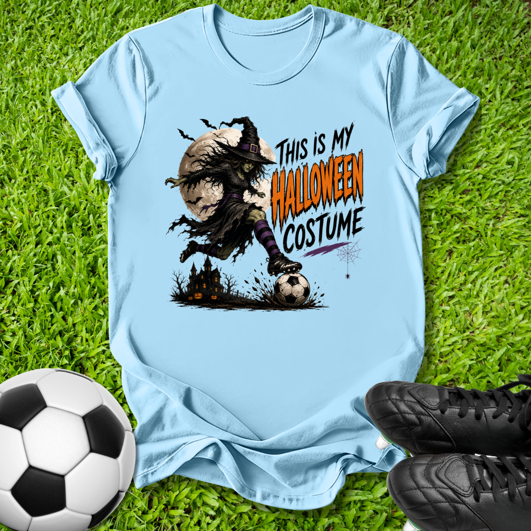 A light blue graphic t-shirt laid flat on green grass, styled with a soccer ball on the left and black cleats on the right. The t-shirt features a graphic of a zombie-like character wearing a Halloween costume kicking a soccer ball, with the text 'THIS IS MY HALLOWEEN COSTUME' printed on the front.