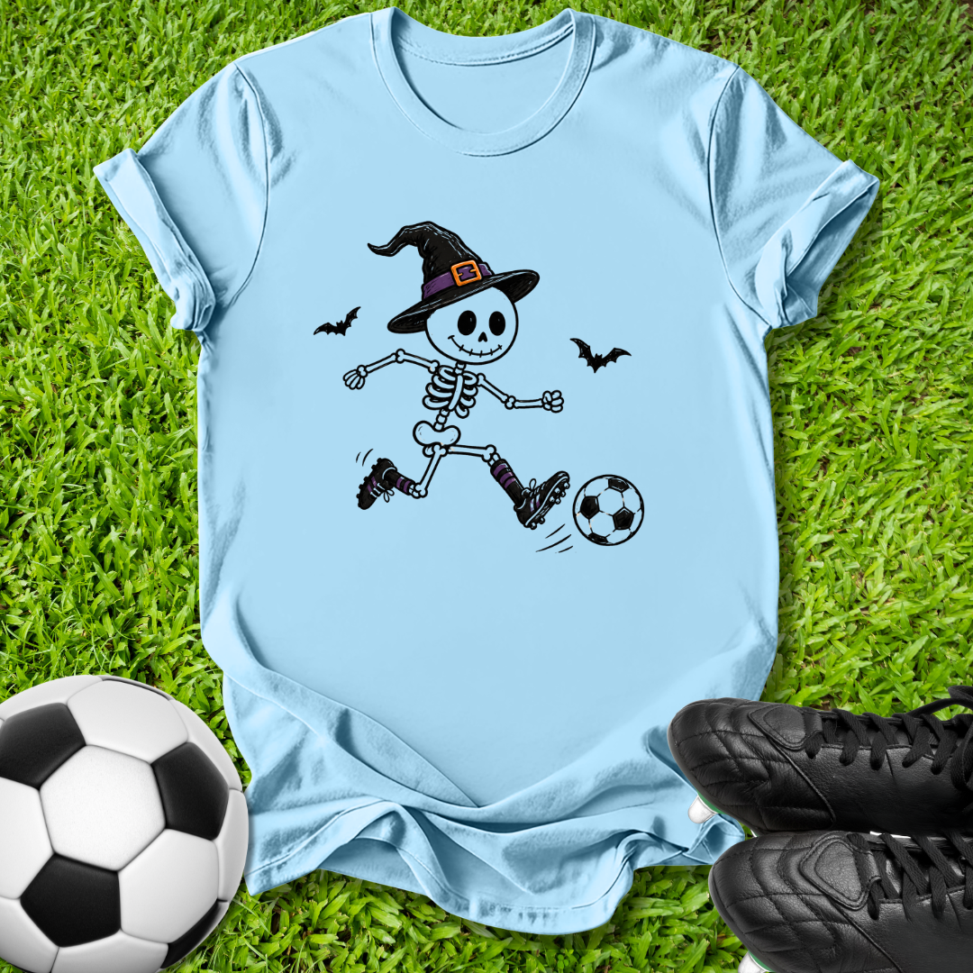 A light blue short-sleeve T-shirt laid flat on green grass, featuring a cartoon skeleton wearing a witch hat and kicking a soccer ball. A real soccer ball and black cleats are placed around it as props.