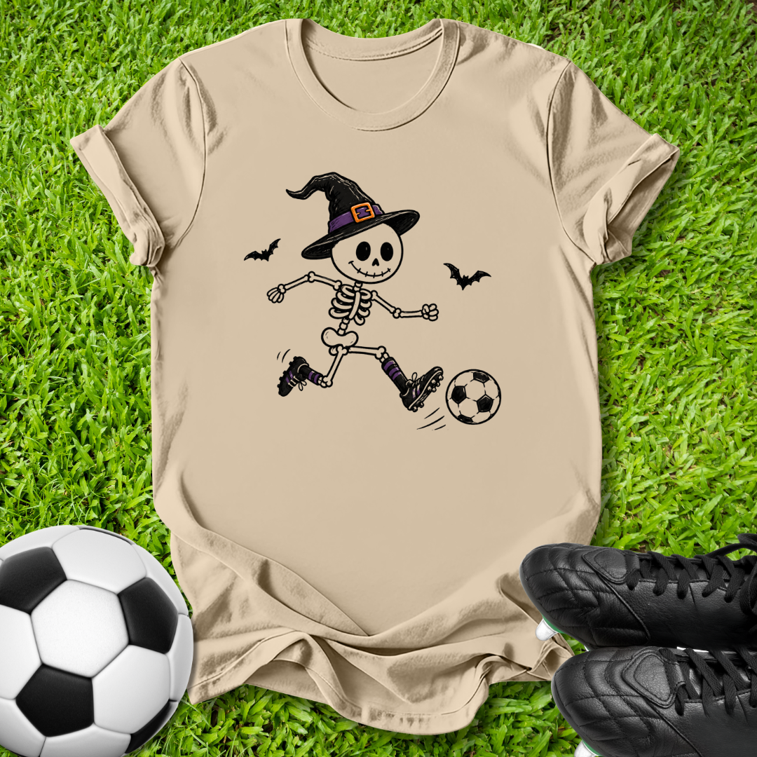 A beige short-sleeve t-shirt laid flat on green grass, featuring a cartoon graphic of a skeleton in a witch hat kicking a soccer ball. A real soccer ball is visible at the bottom left, and black soccer cleats are visible at the bottom right.