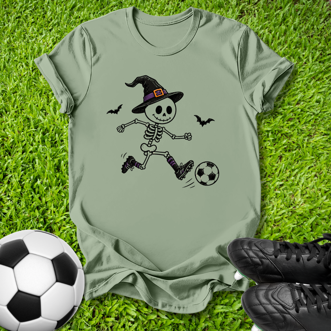 An olive green short-sleeve t-shirt laid flat on green grass, featuring a cartoon graphic of a skeleton in a witch hat kicking a soccer ball. A real soccer ball and black cleats are placed around it as props.