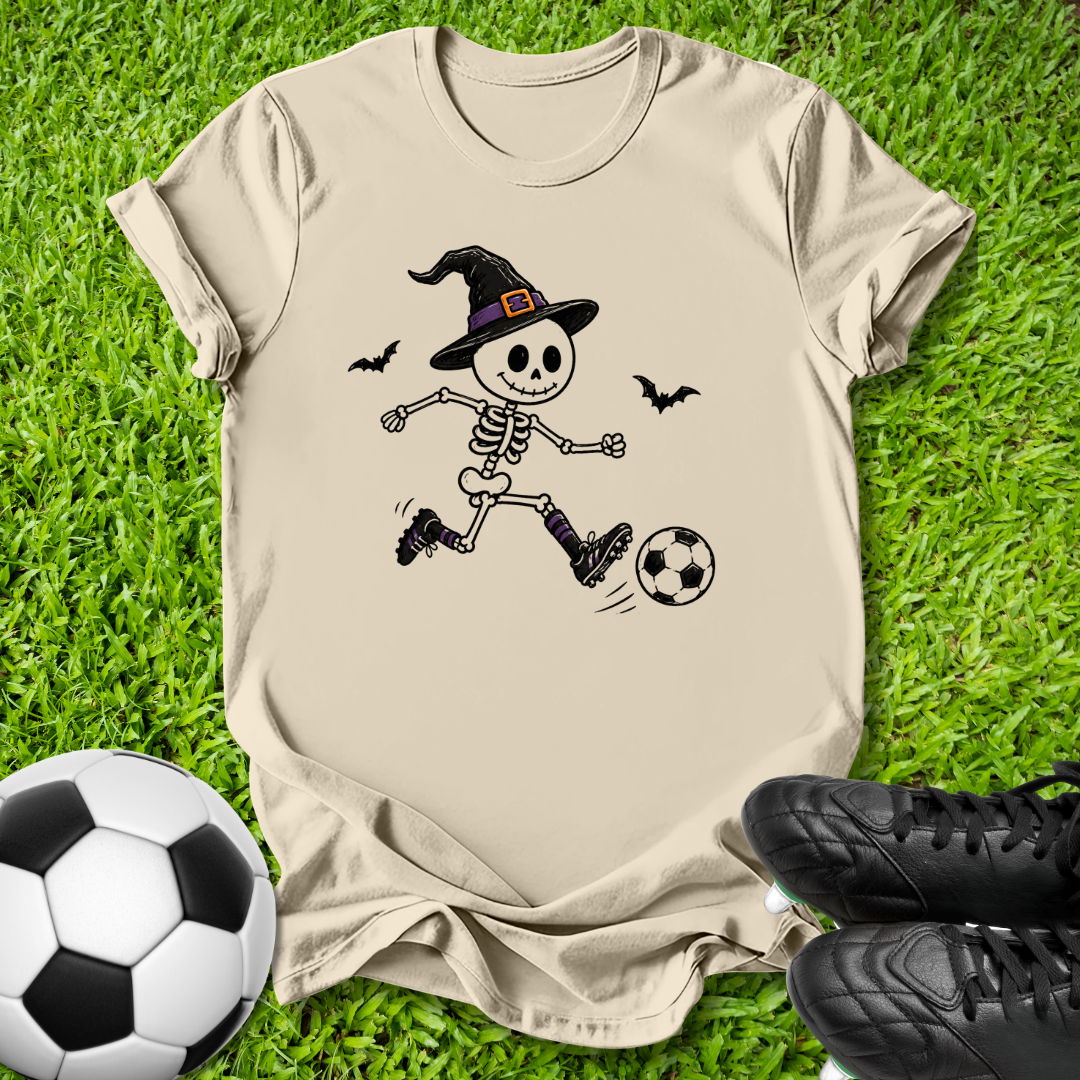 A beige short-sleeve T-shirt laid flat on green grass, featuring a cartoon skeleton in a witch hat kicking a soccer ball. A real soccer ball and black cleats are placed around it as props.
