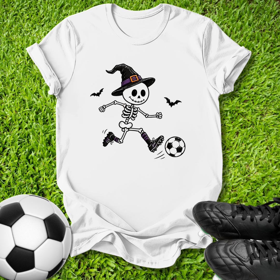 A white short-sleeve T-shirt laid flat on green grass, featuring a cartoon skeleton wearing a witch hat and kicking a soccer ball. A real soccer ball appears at the lower left, and black soccer shoes appear at the lower right, creating a styled outdoor flat-lay scene.