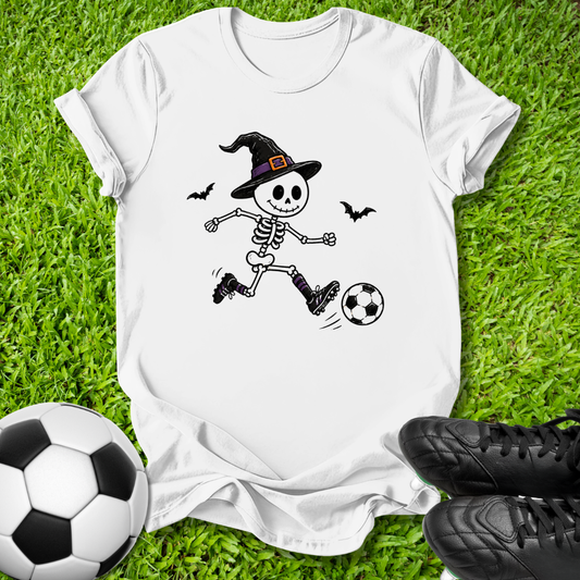 A white short-sleeve T-shirt laid flat on green grass, featuring a cartoon skeleton wearing a witch hat and kicking a soccer ball. A real soccer ball appears at the lower left, and black soccer shoes appear at the lower right, creating a styled outdoor flat-lay scene.