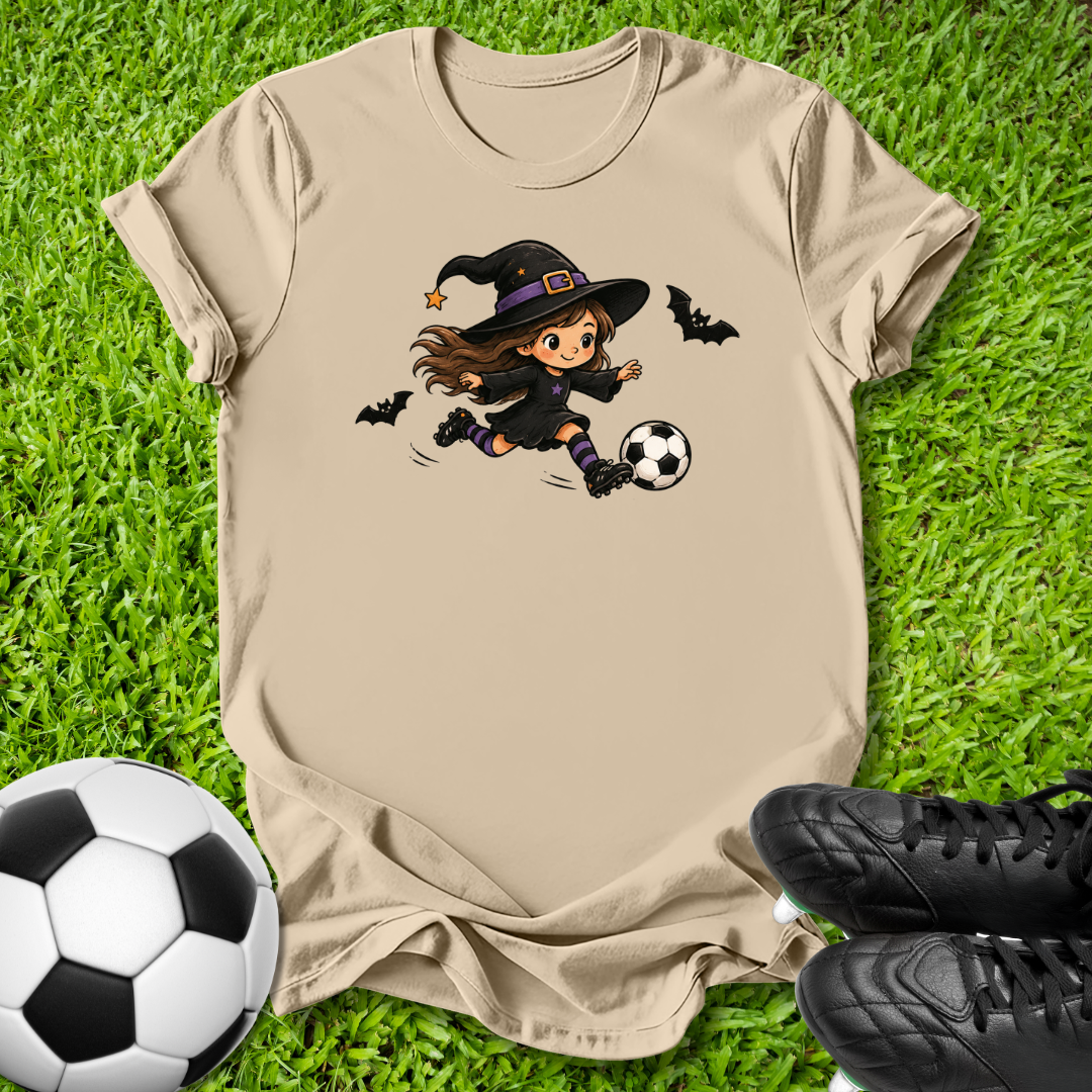 A beige short-sleeve t-shirt laid flat on green grass, featuring a cartoon graphic of a witch in a black hat kicking a soccer ball with bats around her. A soccer ball and black cleats are placed around the shirt as props.