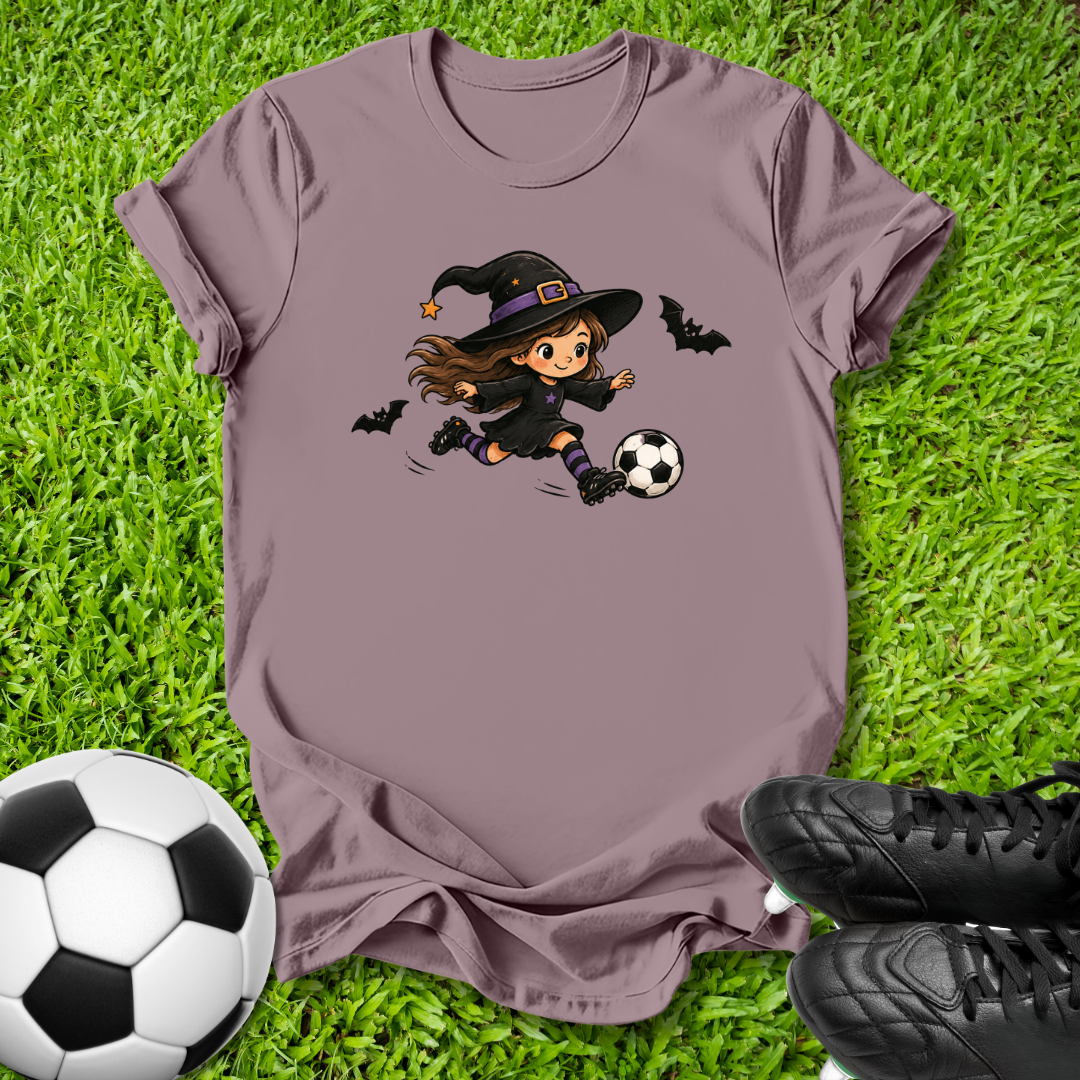 A mauve short-sleeve t-shirt laid flat on green grass, featuring a cartoon graphic of a witch in a black hat kicking a soccer ball, with two black bats flying around. A soccer ball and black cleats are placed around the shirt as props.