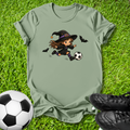 An olive green short-sleeve t-shirt laid flat on green grass, featuring a cartoon graphic of a witch in a black hat kicking a soccer ball, with two small bats. A soccer ball and black cleats are placed around it as props.