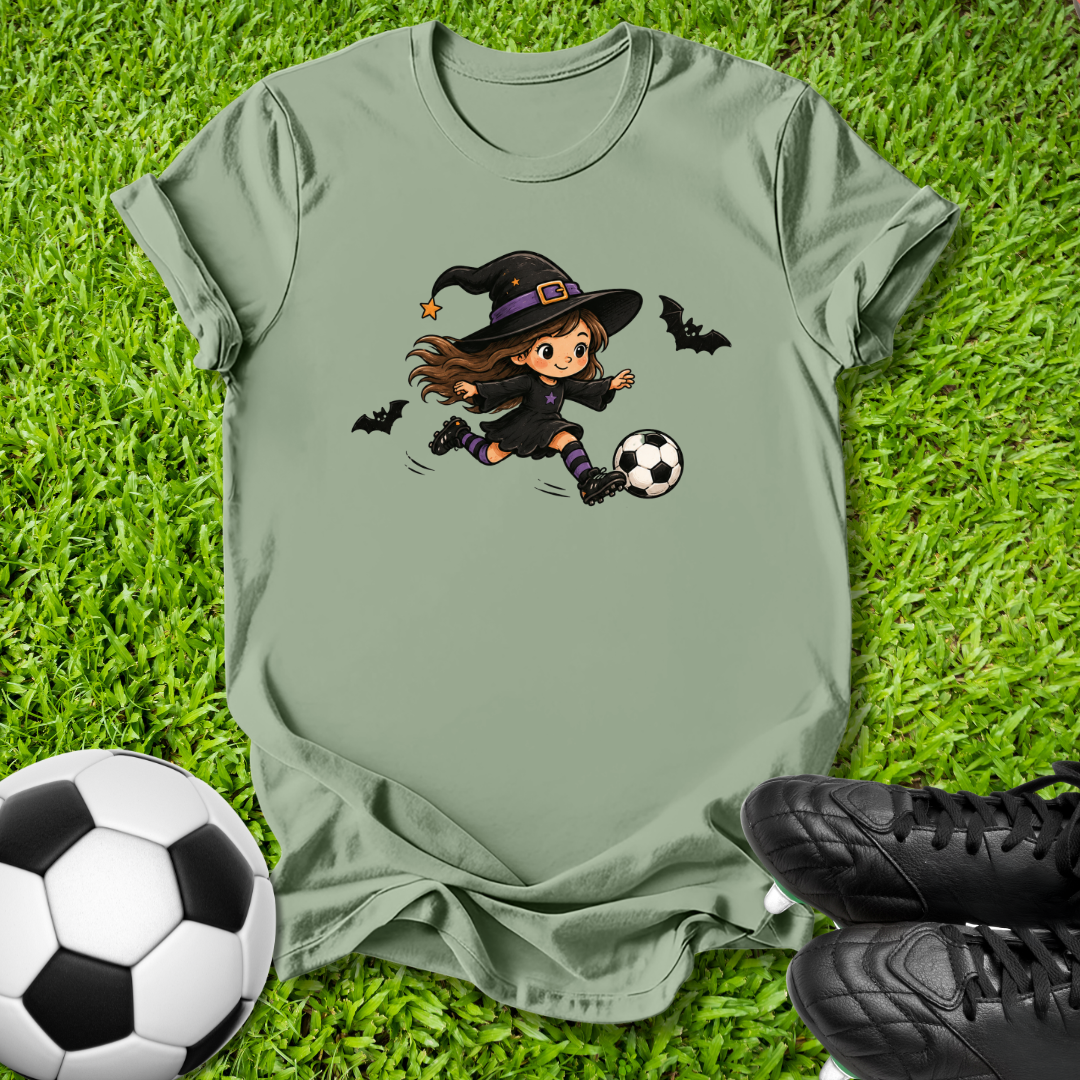 An olive green short-sleeve t-shirt laid flat on green grass, featuring a cartoon graphic of a witch in a black hat kicking a soccer ball, with two small bats. A soccer ball and black cleats are placed around it as props.