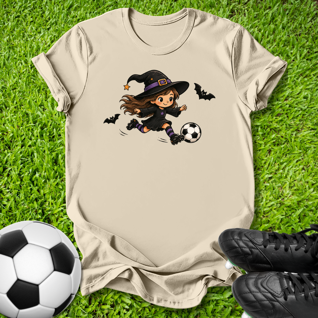 A beige graphic t-shirt laid flat on green grass, featuring a cartoon witch in a black hat kicking a soccer ball with bats flying around. A real soccer ball and black cleats are placed around the shirt as props.