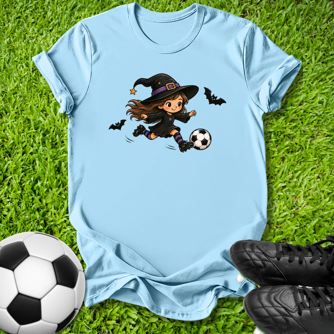 A light blue short-sleeve t-shirt laid flat on green grass, featuring a cartoon graphic of a girl in a witch hat kicking a soccer ball, with two bats flying around. A soccer ball and black cleats are placed around the shirt as props.