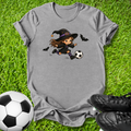 A heather gray short-sleeve t-shirt with a cartoon graphic of a witch kicking a soccer ball, styled on green grass with a soccer ball and black cleats.