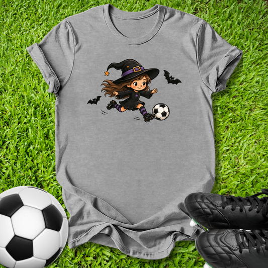 A heather gray short-sleeve t-shirt with a cartoon graphic of a witch kicking a soccer ball, styled on green grass with a soccer ball and black cleats.