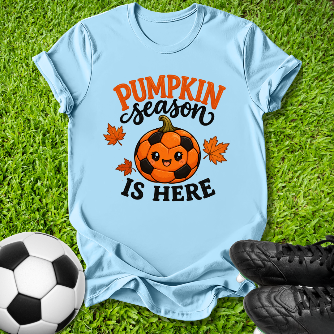 A light blue graphic t-shirt laid flat on green grass, featuring a cute soccer pumpkin design with the text 'PUMPKIN season IS HERE' in orange and black. A soccer ball and black cleats are styled around the shirt.