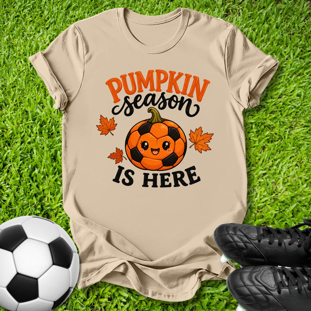 A beige graphic t-shirt laid flat on green grass, featuring a cartoon pumpkin soccer ball and the text 'PUMPKIN season IS HERE' in orange and black. A soccer ball is on the left and black cleats are on the right.