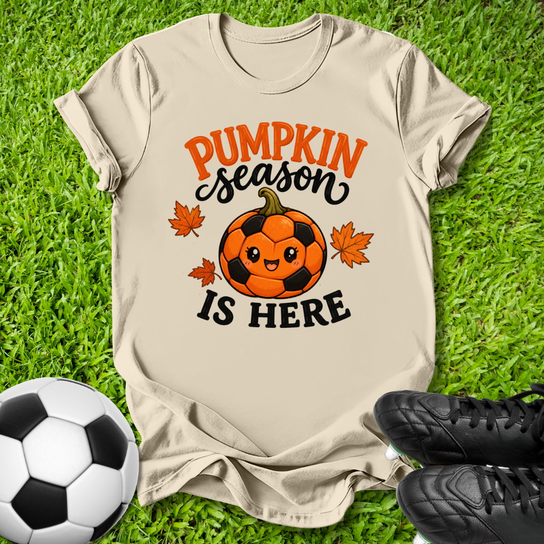 A beige graphic t-shirt laid flat on green grass, featuring a 'Pumpkin Season Is Here' design with a cute soccer pumpkin and orange maple leaves. A soccer ball and black cleats are styled around the shirt.