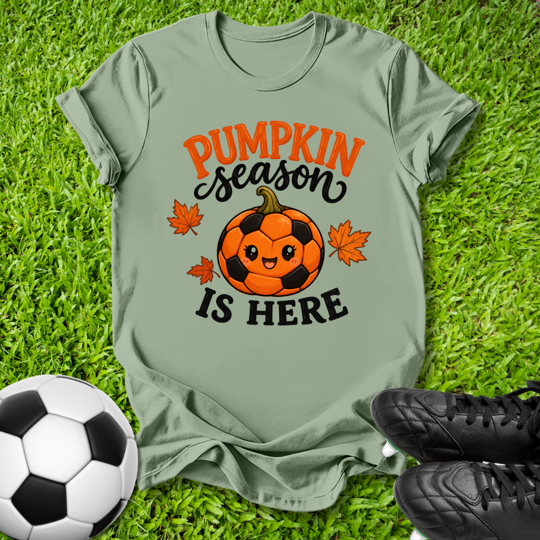An olive green graphic t-shirt laid flat on green grass, featuring a cute soccer pumpkin design with the text 'PUMPKIN season IS HERE' in orange and black. A soccer ball and black cleats are styled around the shirt.