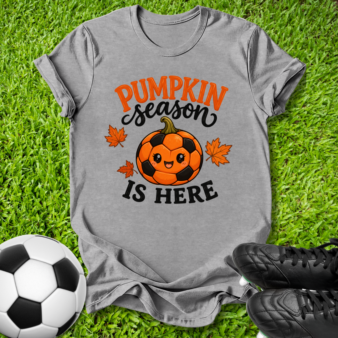 A heather gray graphic t-shirt laid flat on green grass, featuring a cute orange and black soccer pumpkin with the text 'PUMPKIN season IS HERE' and small orange maple leaves. A soccer ball and black cleats are styled around the shirt.