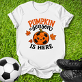 A white graphic t-shirt laid flat on green grass, featuring a 'Pumpkin Season Is Here' design with a soccer pumpkin and maple leaves. A soccer ball is on the left and black cleats are on the right.