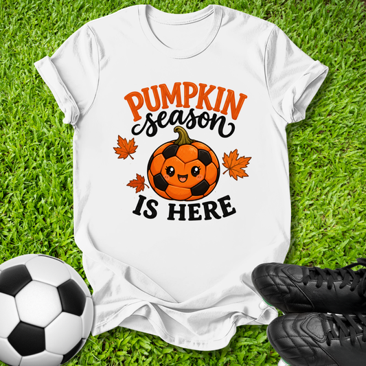 A white graphic t-shirt laid flat on green grass, featuring a 'Pumpkin Season Is Here' design with a soccer pumpkin and maple leaves. A soccer ball is on the left and black cleats are on the right.