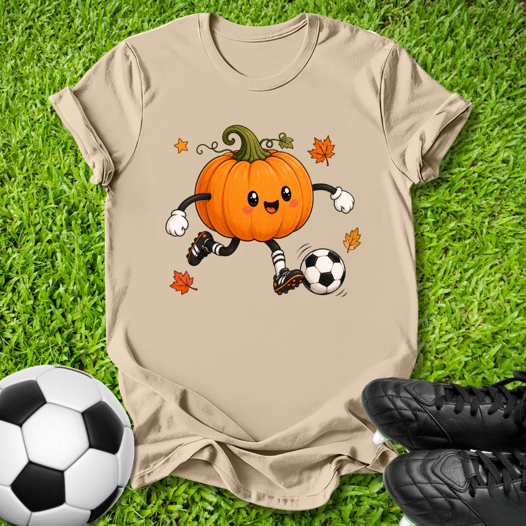 A beige graphic t-shirt laid flat on green grass, featuring a cartoon orange pumpkin character kicking a soccer ball with autumn leaves. A soccer ball and black cleats are placed around it as props.
