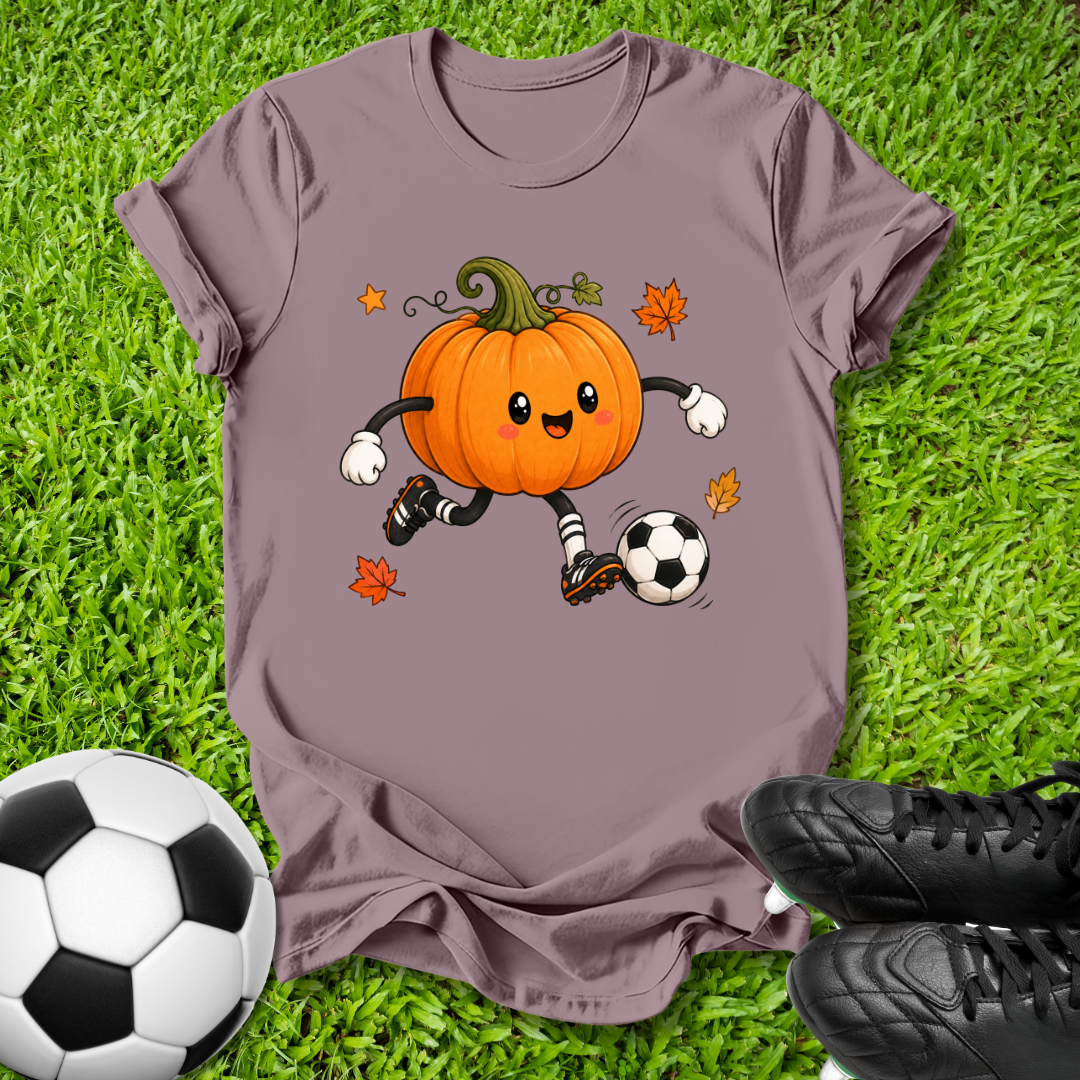 A mauve short-sleeve t-shirt laid flat on green grass, featuring a cartoon orange pumpkin character kicking a soccer ball with small autumn leaves around it. A real soccer ball and black cleats are placed around the shirt as styling props.