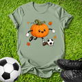 An olive green short-sleeve t-shirt laid flat on green grass, featuring a cartoon graphic of an orange pumpkin character soccer-kicking a black and white soccer ball with small orange leaves. A real soccer ball is visible in the bottom-left corner and a pair of black cleats is visible in the bottom-right corner.