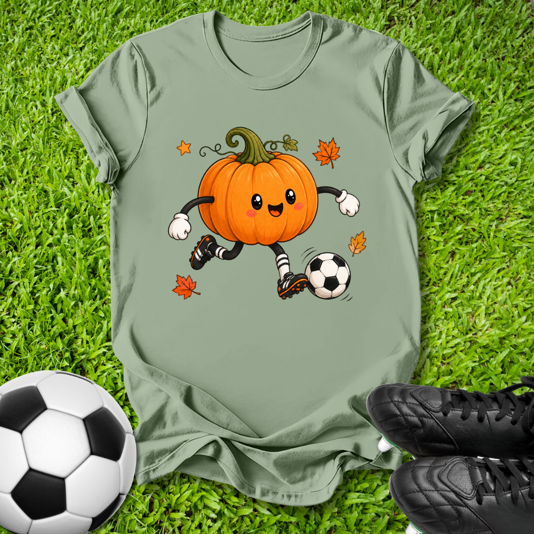 An olive green short-sleeve t-shirt laid flat on green grass, featuring a cartoon graphic of an orange pumpkin character soccer-kicking a black and white soccer ball with small orange leaves. A real soccer ball is visible in the bottom-left corner and a pair of black cleats is visible in the bottom-right corner.