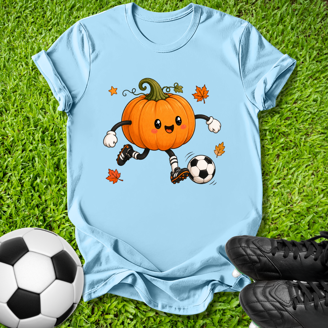 A light blue short-sleeve t-shirt laid flat on green grass, featuring a cartoon graphic of an orange pumpkin character soccer-kicking a black and white ball with small orange leaves. A real soccer ball and black cleats are placed around it as props.