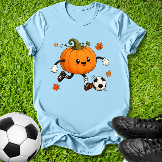 A light blue short-sleeve t-shirt laid flat on green grass, featuring a cartoon graphic of an orange pumpkin character soccer-kicking a black and white ball with small orange leaves. A real soccer ball and black cleats are placed around it as props.