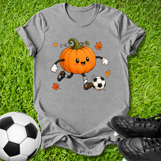 A heather gray short-sleeve t-shirt laid flat on green grass, featuring a cartoon graphic of an orange pumpkin character soccer-kicking a black and white soccer ball with small orange leaves around it. A real soccer ball and black cleats are placed as props around the shirt.