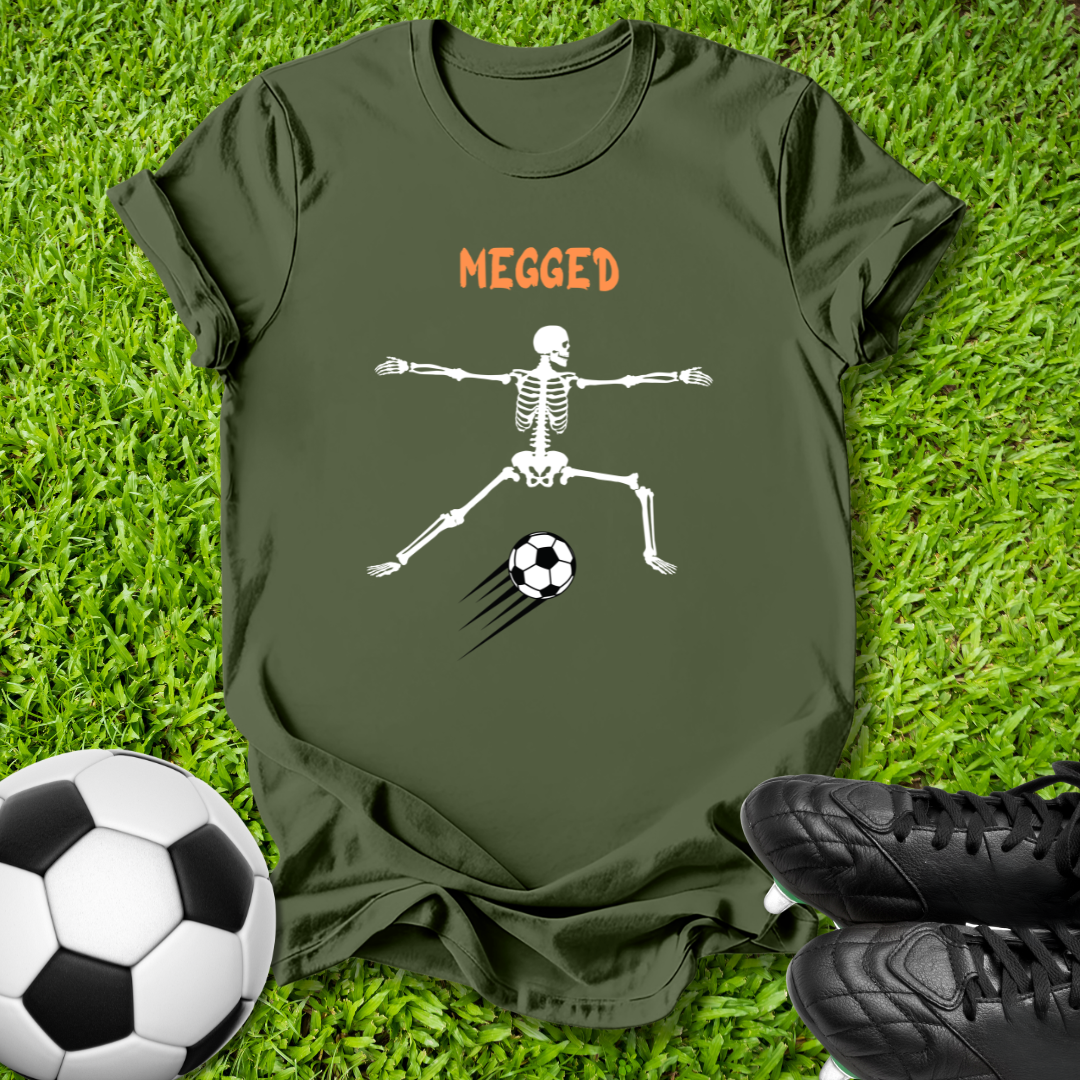 An olive green t-shirt laid flat on green grass, featuring a white skeleton graphic kicking a soccer ball with the word 'MEGGED' in orange above. A soccer ball and black cleats are placed around the shirt.