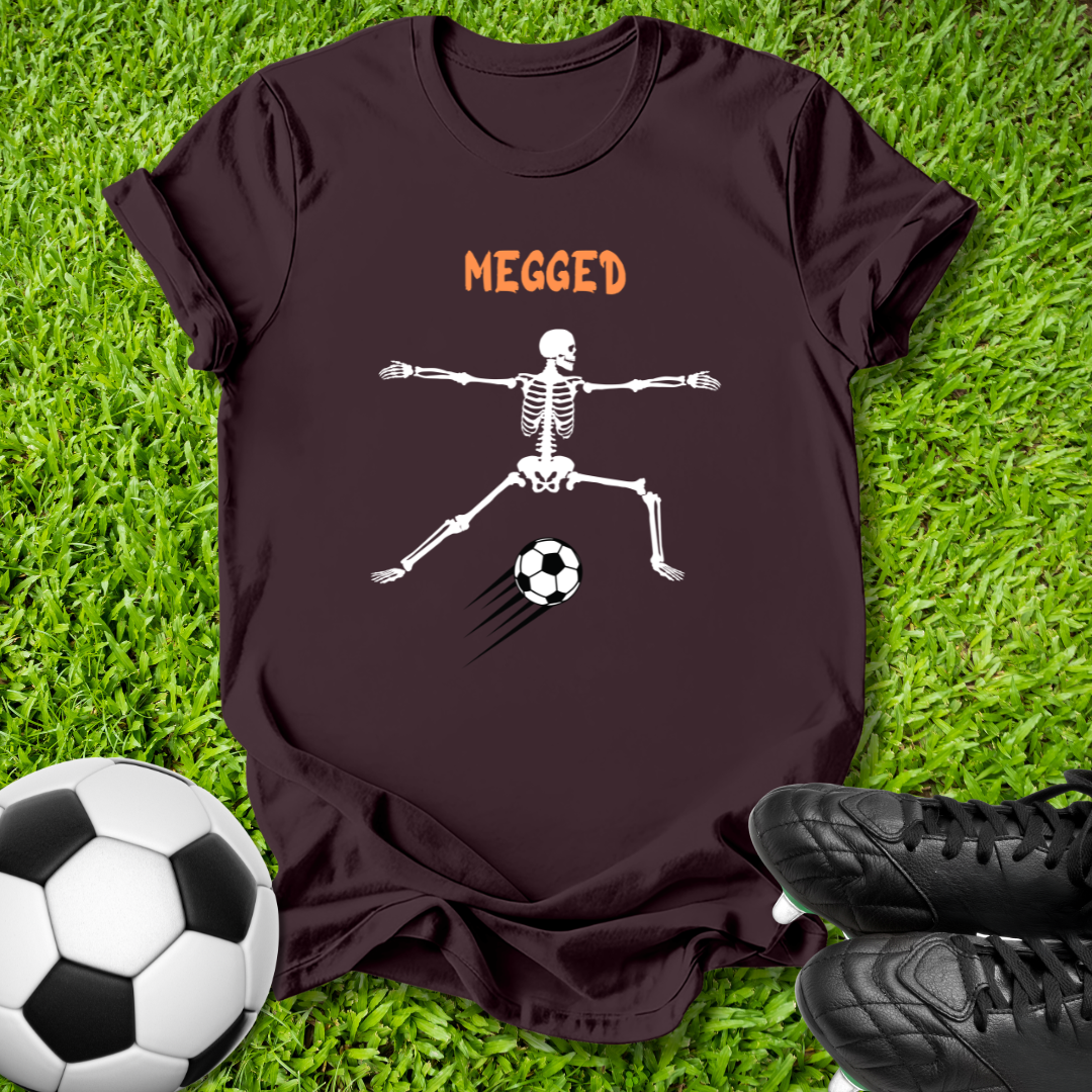 A dark brown t-shirt laid flat on green grass, featuring a white skeleton graphic kicking a soccer ball with the word 'MEGGED' in orange above. A soccer ball and black cleats are styled around the shirt.