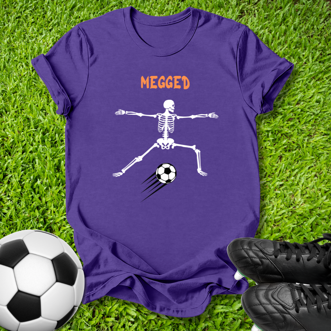 A purple short-sleeve t-shirt laid flat on green grass, featuring a white skeleton graphic kicking a black-and-white soccer ball with the word 'MEGGED' in orange above. A soccer ball and black cleats are placed around it as props.