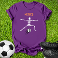 A purple short-sleeve t-shirt laid flat on green grass, featuring a white skeleton kicking a soccer ball with the word 'MEGGED' in orange above. A soccer ball and black cleats are placed around it as props.