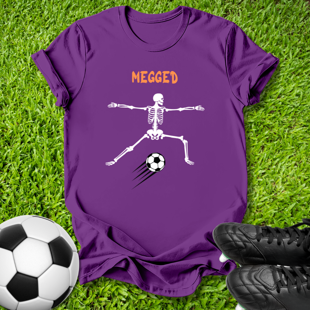 A purple short-sleeve t-shirt laid flat on green grass, featuring a white skeleton kicking a soccer ball with the word 'MEGGED' in orange above. A soccer ball and black cleats are placed around it as props.
