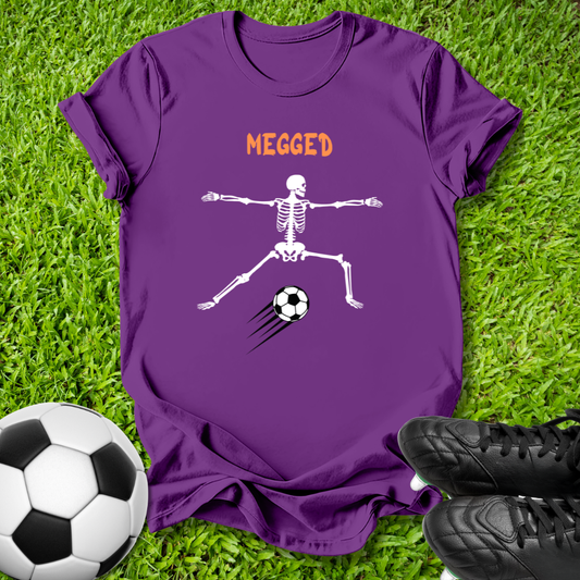 A purple short-sleeve t-shirt laid flat on green grass, featuring a white skeleton kicking a soccer ball with the word 'MEGGED' in orange above. A soccer ball and black cleats are placed around it as props.